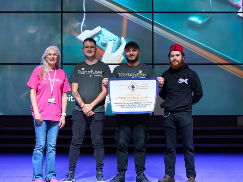Lewis, Ryan and Garrett on stage at ProtoPlay 2025 holding the Tranzfuser Public Vote Winner certificate, beside a ProtoPlay organiser.